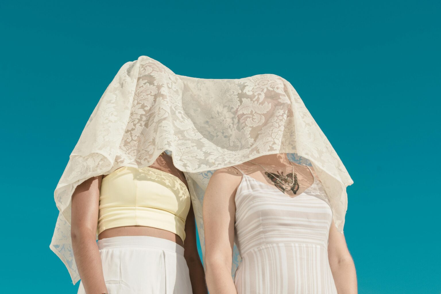Minimalist shot of two women under a white lace veil against a clear blue sky.