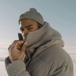 A man wearing winter fashion poses confidently in a snow-covered landscape, showcasing style and warmth.